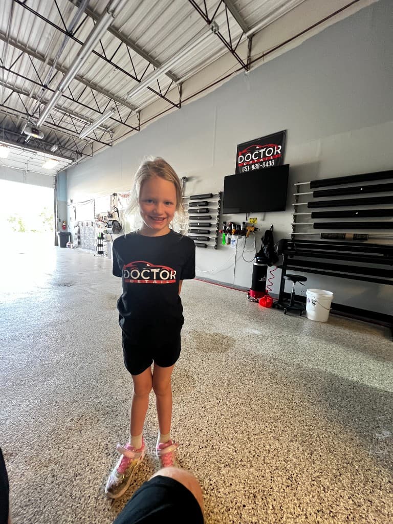 Alice smiling in the Doctor Detail Eagan MN shop — wearing a Doctor Detail shirt with tint film rolls, signage, and epoxy floor visible