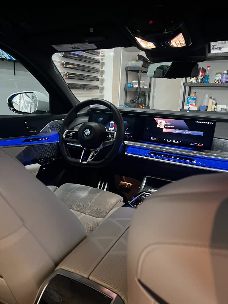 BMW interior from rear seat — curved display, blue ambient lighting, beige leather, shop visible through windows — Doctor Detail Eagan MN